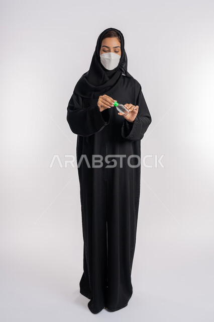 اتباع الاجراءات الاحترازية للوقاية من فايروس كورونا، بورتريه لإمرأة عربية خليجية إماراتية ترتدي العباءة والحجاب تضع الكمامة تمسك علبة المعقم, شابة سعودية تقوم باستخدام المواد الكيميائية والكحول للقضاء على الفايروسات والجراثيم، صورة الجسم بالكامل، خلفية بيضاء
