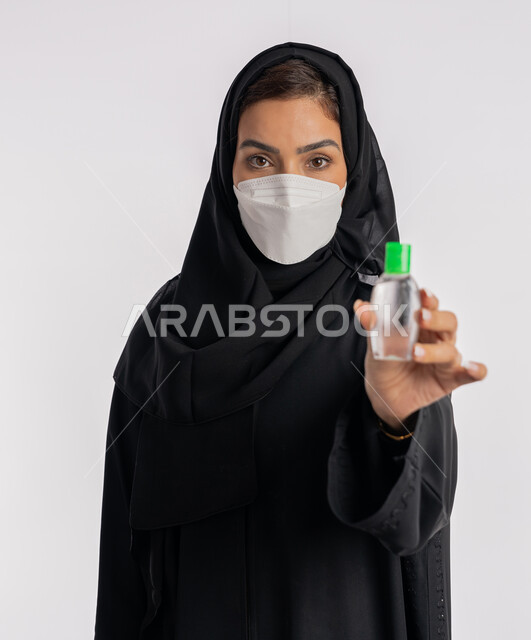 استخدام المواد الكيميائية والكحول للقضاء على الفايروسات والجراثيم، شابة سعودية اتباع الاجراءات الاحترازية للوقاية من فايروس كورونا، بورتريه مقرب لإمرأة عربية خليجية إماراتية ترتدي العباءة والحجاب تضع الكمامة تمسك علبة المعقم بيدها, خلفية بيضاء
