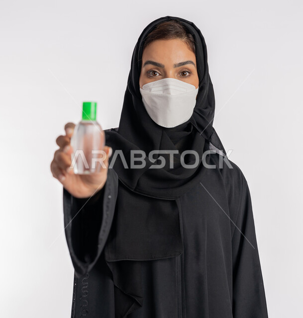 شابة سعودية تتبع الاجراءات الاحترازية للوقاية من فايروس كورونا، بورتريه مقرب لإمرأة عربية خليجية إماراتية ترتدي العباءة والحجاب تضع الكمامة تمسك علبة المعقم بيدها, استخدام المواد الكيميائية والكحول للقضاء على الفايروسات والجراثيم، خلفية بيضاء
