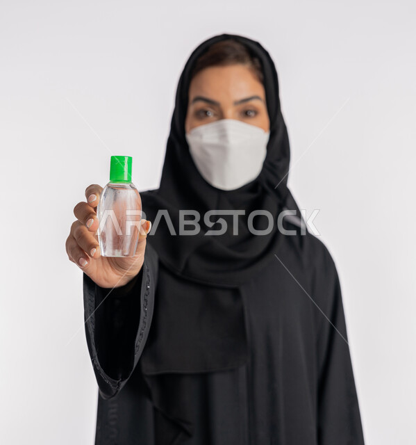 شابة سعودية تتبع الاجراءات الاحترازية للوقاية من فايروس كورونا، بورتريه مقرب لإمرأة عربية خليجية إماراتية ترتدي العباءة والحجاب تضع الكمامة تمسك علبة المعقم بيدها, استخدام المواد الكيميائية والكحول للقضاء على الفايروسات والجراثيم، خلفية بيضاء