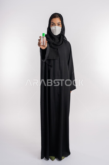 اتباع الاجراءات الاحترازية للوقاية من فايروس كورونا، بورتريه لإمرأة عربية خليجية إماراتية ترتدي العباءة والحجاب تضع الكمامة تمسك علبة المعقم, شابة سعودية تقوم باستخدام المواد الكيميائية والكحول للقضاء على الفايروسات والجراثيم، صورة الجسم بالكامل، خلفية بيضاء