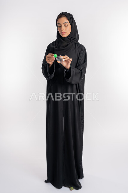 الاهتمام بالنظافة واتباع الاجراءات الوقائية من فايروس كورونا، بورتريه لإمرأة عربية خليجية إماراتية ترتدي العباءة والحجاب تمسك بيدها علبة المعقم, شابة سعودية تقوم باستخدام المواد الكيميائية والكحول للقضاء على الفايروسات والجراثيم، صورة الجسم بالكامل، خلفية بيضاء