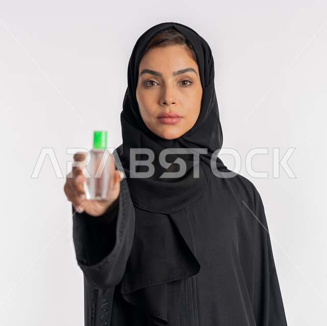 Using chemicals and alcohol to eliminate viruses and germs, taking care of cleanliness and following preventive measures against the Corona virus, close-up portrait of an Arab Gulf Emirati woman wearing an abaya and hijab holding a bottle of sanitizer in her hand, a young Saudi woman sanitizing her hands, white background