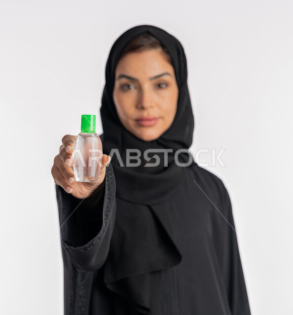 Using chemicals and alcohol to eliminate viruses and germs, taking care of cleanliness and following preventive measures against the Corona virus, close-up portrait of an Arab Gulf Emirati woman wearing an abaya and hijab holding a bottle of sanitizer in her hand, a young Saudi woman sanitizing her hands, white background