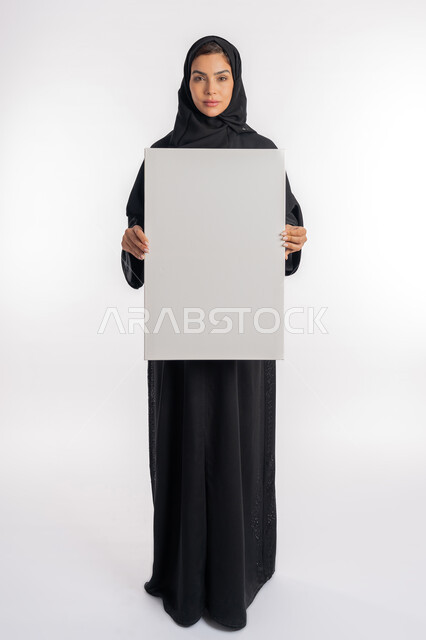 لوحات توضيحية ووسائل تعليمية, شابة سعودية تنظر إلى الكاميرا بتعابير الحزم والجدية، العروض والإعلانات التسويقية, بورتريه لإمرأة عربية خليجية إماراتية ترتدي الحجاب والعباءة تمسك بيديها لوحة بيضاء مستطيلة فارغة، موك اب مستطيل أبيض, صورة الجسم بالكامل، خلفية بيضاء