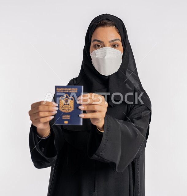 اتباع الاجراءات الاحترازية للوقاية من فايروس كورونا في السفر، بورتريه مقرب لإمرأة عربية خليجية إماراتية محجبة ترتدي الكمامة تمسك بيدها باسبور الامارات, الوعي الصحي لتجنب إنتشار العدوى، الحفاظ على السلامة من كوفيد 19، شابة سعودي تحمل جواز السفر، خلفية بيضاء