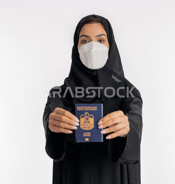 الوعي الصحي لتجنب إنتشار العدوى، الحفاظ على السلامة من كوفيد 19، شابة سعودي تحمل جواز السفر، اتباع الاجراءات الاحترازية للوقاية من فايروس كورونا في السفر، بورتريه مقرب لإمرأة عربية خليجية إماراتية محجبة ترتدي الكمامة تمسك بيدها باسبور الامارات, خلفية بيضاء