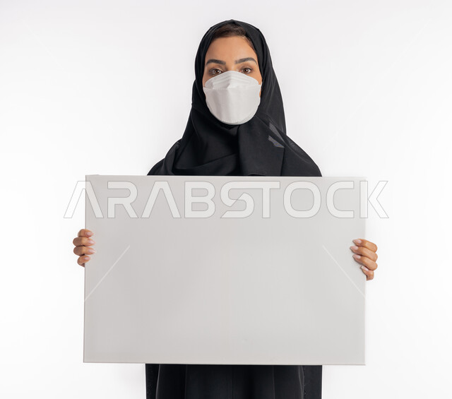 لوحات توعوية لتجنب إنتشار العدوى، الحفاظ على السلامة من كوفيد 19، شابة سعودية تقوم باتباع الاجراءات الاحترازية للوقاية من فايروس كورونا، بورتريه مقرب لإمرأة عربية خليجية إماراتية محجبة ترتدي الكمامة تمسك بيدها موك اب مستطيل أبيض فارغ, خلفية بيضاء