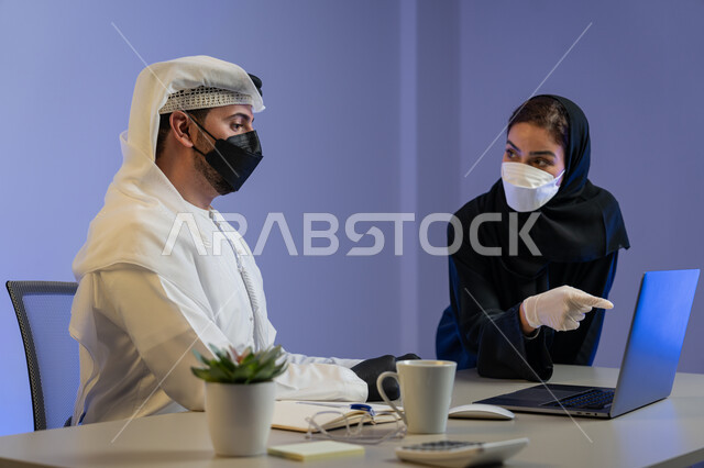 تبادل الخبرات بين الموظفين لتحقيق اهداف الشركة، ارتداء الكمامة الطبية والقفازات لمنع انتشار فايروس كورونا، صورة مقربة لزميليين عربيين خليجيين إماراتيين يقومان بإنجاز الاعمال باستخدام الحاسب المحمول، اتباع الاجراءات الوقائية من الأمراض في الشركات الاماراتية