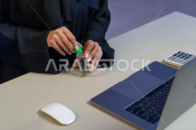 الاهتمام بالنظافة واتباع الاجراءات الوقائية من فايروس كورونا، صورة مقربة لإمرأة عربية خليجية إماراتية تجلس امام الحاسوب المحمول تقوم بتعقيم يديها, شابة سعودية تستخدم المواد الكيميائية والكحول للقضاء على الفايروسات، مهن ووظائف نسائية، استخدام الاجهزة التقنية الحديثة