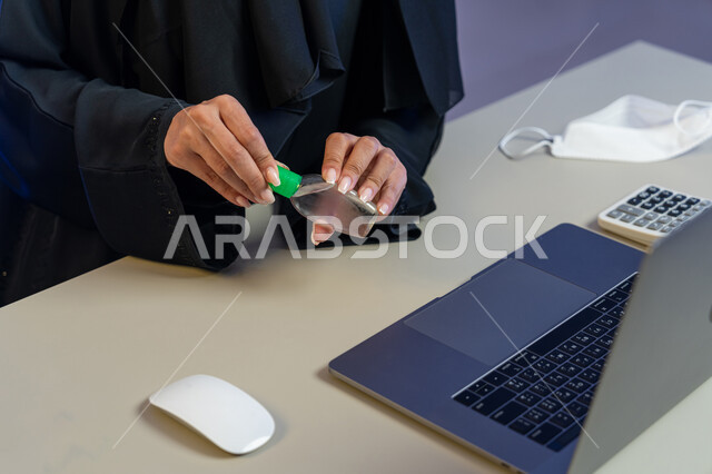 مهن ووظائف نسائية، استخدام الاجهزة التقنية الحديثة، الاهتمام بالنظافة واتباع الاجراءات الوقائية من فايروس كورونا، صورة مقربة لإمرأة عربية خليجية إماراتية تجلس امام الحاسوب المحمول تقوم بتعقيم يديها, شابة سعودية تستخدم المواد الكيميائية والكحول للقضاء على الفايروسات