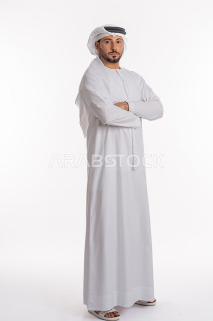 مفهوم الرجولة والثقة بالنفس، الأناقة والاهتمام بالمظهر الخارجي, بورتريه لشاب عربي خليجي إماراتي أنيق يرتدي الكندورة والغترة يقف بيدين متقاطعتين ينظر إلى الكاميرا بإيماءات السعادة والفرح, صورة الجسم بالكامل، خلفية بيضاء