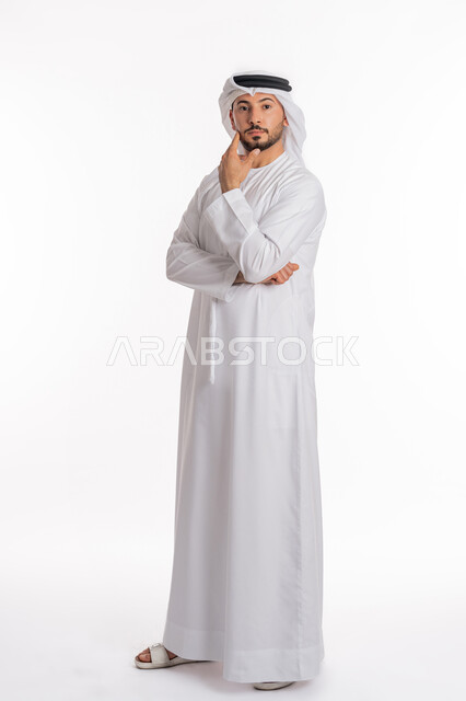Planning for the future and setting future goals, looking at the camera with absentminded expressions, portrait of a young Arab Gulf Emirati man wearing a kandura and ghutra standing and putting his hand on his chin with gestures of worry and thinking about something, full body photo, white background