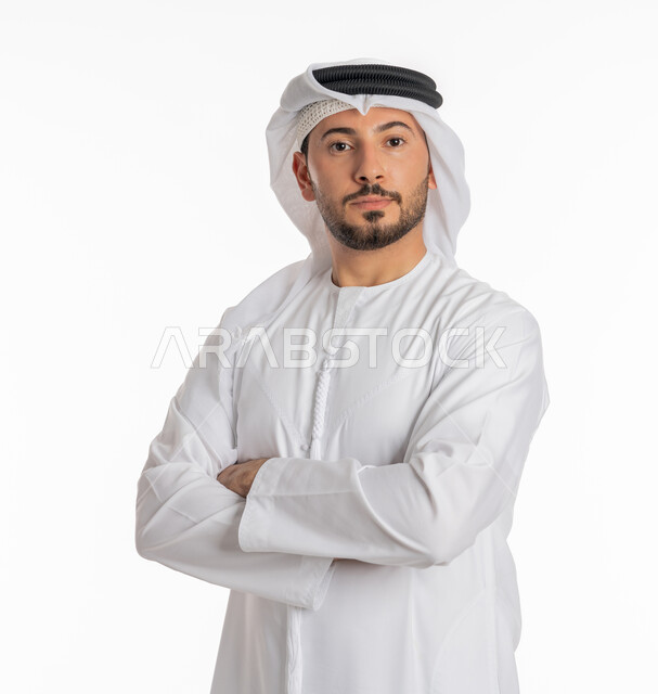 الأناقة والاهتمام بالمظهر الخارجي, بورتريه مقرب لشاب عربي خليجي إماراتي أنيق يرتدي الكندورة والغترة يقف بيدين متقاطعتين ينظر إلى الكاميرا بإيماءات السعادة والفرح, مفهوم الرجولة والثقة بالنفس، خلفية بيضاء