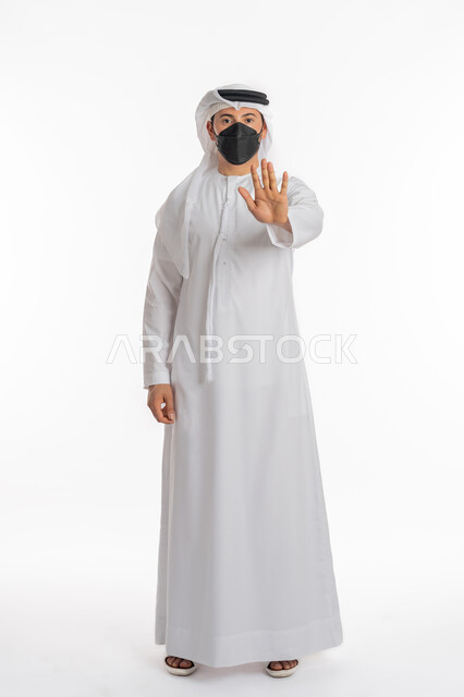 التحذير وطلب التوقف, تجنب انتشار العدوى بكوفيد 19، اتباع الإجراءات الوقائية من فايروس كورونا، بورتريه لرجل عربي إماراتي خليجي يرتدي الكندورة والغترة يضع الكمامة يرفع كف يده لأعلى بتعابير الرفض ومنع الاقتراب للحفاظ على السلامة, صورة الجسم بالكامل، خلفية بيضاء