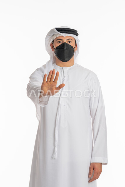 تجنب انتشار العدوى بكوفيد 19، الرفض ومنع الاقتراب للحفاظ على السلامة, اتباع الإجراءات الاحترازية للوقاية من فايروس كورونا، بورتريه لرجل عربي إماراتي خليجي يرتدي الكندورة والغترة يضع الكمامة يرفع كف يده لأعلى بتعابير التحذير والتوقف, خلفية بيضاء