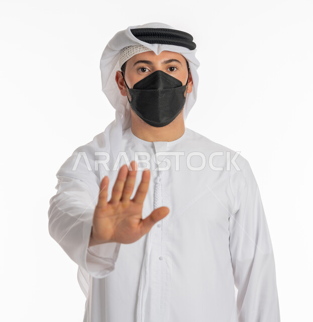 الرفض ومنع الاقتراب للحفاظ على السلامة, تجنب انتشار العدوى بكوفيد 19، اتباع الإجراءات الاحترازية للوقاية من فايروس كورونا، بورتريه مقرب لرجل عربي إماراتي خليجي يرتدي الكندورة والغترة يضع الكمامة يرفع كف يده لأعلى بتعابير التحذير والتوقف, خلفية بيضاء