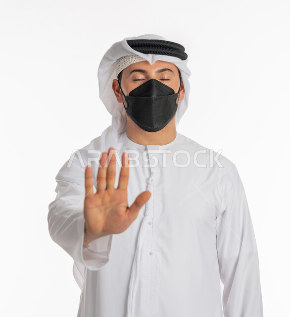 Refusal and prevention of approach to maintain safety, avoid the spread of Covid-19 infection, follow precautionary measures to prevent the Corona virus, close-up portrait of an Emirati Gulf Arab man wearing a kandura and a ghutra, wearing a mask, raising his palm up with warning and stop expressions, white background