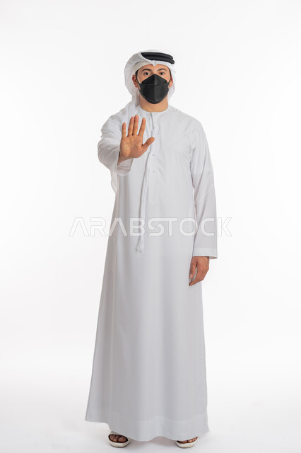 التحذير وطلب التوقف, تجنب انتشار العدوى بكوفيد 19، اتباع الإجراءات الوقائية من فايروس كورونا، بورتريه لرجل عربي إماراتي خليجي يرتدي الكندورة والغترة يضع الكمامة يرفع كف يده لأعلى بتعابير الرفض ومنع الاقتراب للحفاظ على السلامة, صورة الجسم بالكامل، خلفية بيضاء