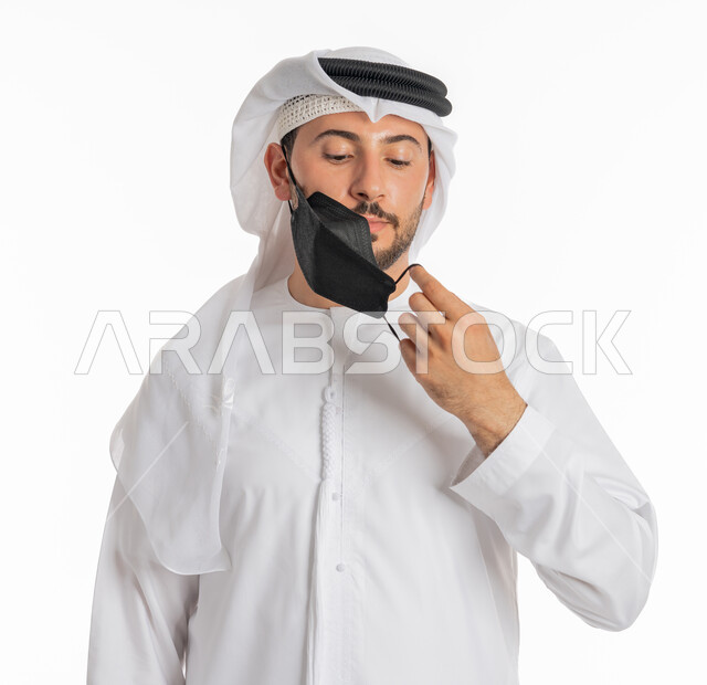 ارتداء القناع لإيقاف عدوى كوفيد 19, الحيطة من انتقال المرض, اتباع إجراءات السلامة للوقاية من العدوى, توخي الحذر من انتشار فيروس كورونا, بورتريه مقرب لرجل عربي خليجي إماراتي يرتدي الكندورة والغترة يقوم بارتداء الكمامة الطبية, خلفية بيضاء