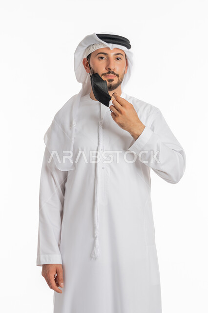 الحيطة من انتقال المرض, ارتداء القناع لإيقاف عدوى كوفيد 19, اتباع إجراءات السلامة للوقاية من العدوى, توخي الحذر من انتشار فيروس كورونا, بورتريه مقرب لرجل عربي خليجي إماراتي يرتدي الكندورة والغترة يقوم بارتداء الكمامة الطبية, خلفية بيضاء