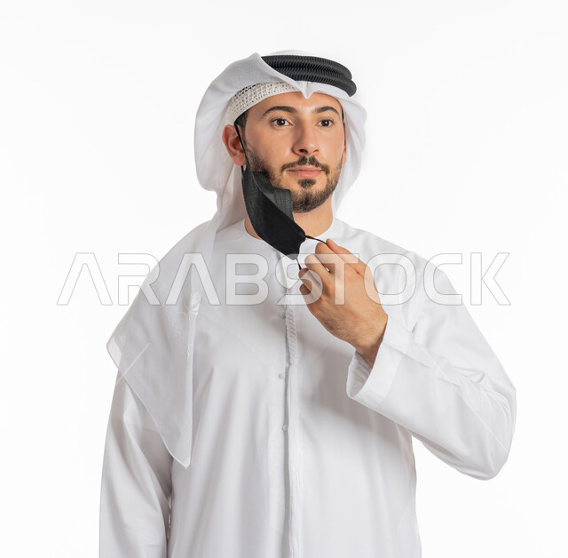 الحيطة من انتقال المرض, ارتداء القناع لإيقاف عدوى كوفيد 19, اتباع إجراءات السلامة للوقاية من العدوى, توخي الحذر من انتشار فيروس كورونا, بورتريه مقرب لرجل عربي خليجي إماراتي يرتدي الكندورة والغترة يقوم بارتداء الكمامة الطبية, خلفية بيضاء