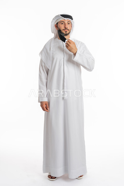 Following safety measures to prevent infection, Being careful of the spread of the Corona virus, Portrait of an Emirati Gulf Arab man wearing a kandura and ghutra wearing a medical mask, Wearing a mask to stop Covid-19 infection, Being careful of the transmission of the disease, Full body image, White background
