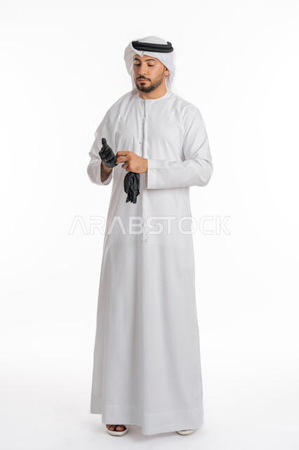 اتباع إجراءات السلامة للوقاية من العدوى, توخي الحذر من انتشار فيروس كورونا, بورتريه لرجل عربي خليجي إماراتي يرتدي الكندورة والغترة يقوم بارتداء القفازات, الحيطة من انتقال المرض, منع وتجنب ملامسة الايدي للجراثيم, صورة الجسم بالكامل، خلفية بيضاء