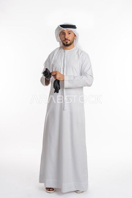اتباع إجراءات السلامة للوقاية من العدوى, توخي الحذر من انتشار فيروس كورونا, بورتريه لرجل عربي خليجي إماراتي يرتدي الكندورة والغترة يقوم بارتداء القفازات, الحيطة من انتقال المرض, منع وتجنب ملامسة الايدي للجراثيم, صورة الجسم بالكامل، خلفية بيضاء