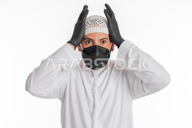 تعابير الاستياء والضجر من المرض، اتباع الاجراءات الاحترازية للوقاية من فايروس كورونا، الصداع وألم الرأس والمعاناة من آلام الشقيقة، بورتريه مقرب لرجل عربي خليجي إماراتي يرتدي الكندورة والغترة يضع الكمامة يرفع يديه على رأسه بايماءات الارهاق، خلفية بيضاء