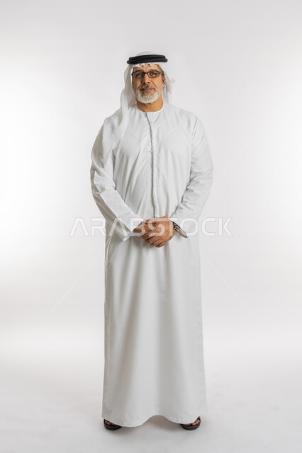 The concept of caring about external appearance, masculinity and elegance, gestures of self-confidence, looking at the camera, gestures of pleasure, a portrait of an elderly Arab, Gulf, Emirati man wearing a kandora and a ghutra, standing straight with knit hand gestures, full-length body portrait, white background.