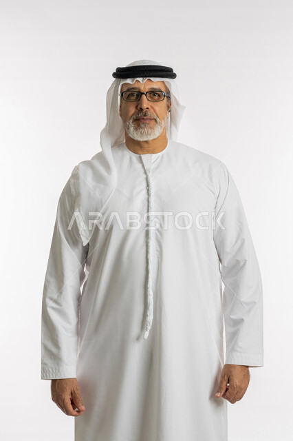النظر إلى الكاميرا، مفهوم الرجولة والأناقه ,تعابير الجدية والثقة بالنفس، الاهتمام بالمظهر الخارجي، بورتريه لرجل مسن عربي خليجي اماراتي يرتدي الكندورة و الغترة يقف باستقامة ,خلفية بيضاء