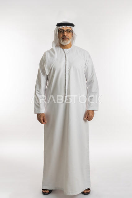 مفهوم  الرجولة والأناقه ,تعابير الجدية والثقة بالنفس، الاهتمام بالمظهر الخارجي، بورتريه لرجل مسن عربي خليجي اماراتي يرتدي الكندورة و الغترة يقف باستقامة ,النظر إلى الكاميرا، صورة الجسم بالطول الكامل، خلفية بيضاء