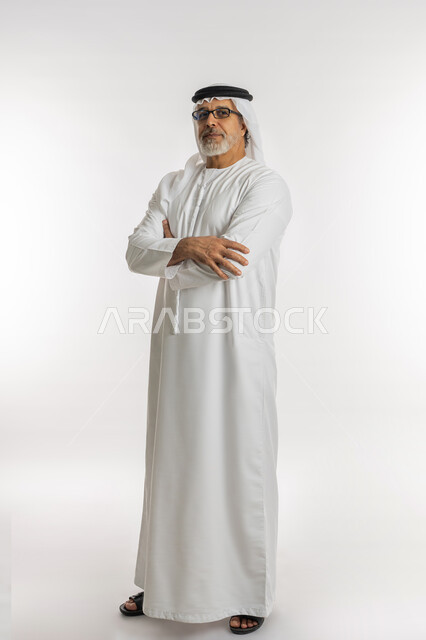 النظر إلى الكاميرا يإيماءات السرور، ايماءات الثقة بالنفس، مفهوم الاهتمام بالمظهر الخارجي و الرجولة والأناقه، بورتريه مقرب لرجل مسن عربي خليجي اماراتي يرتدي الكندورة و الغترة يقف باستقامة بايماءات ايدي متقاطعه ,خلفية بيضاء