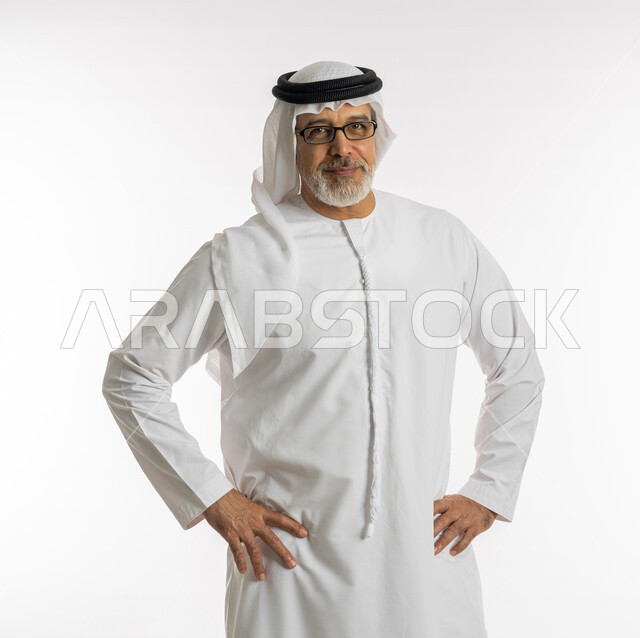 الوقوف بتعابير تدل على الانتظار، الملل والشرود والتفكير، بورتريه لرجل كبير السن عربي اماراتي خليجي يرتدي الغترة و الكندورة و النظارات الطبية يضع يديه على خصره و ينظر الى الكاميرا بايماءات السرور ،خلفية بيضاء