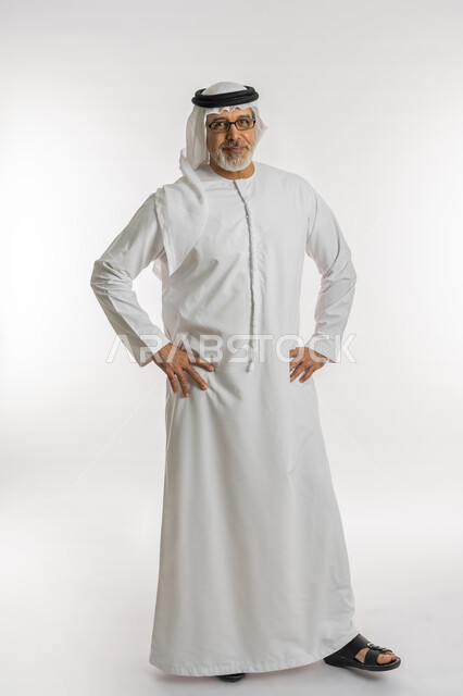 الملل والشرود والتفكير، بورتريه لرجل كبير السن عربي اماراتي خليجي يرتدي الغترة و الكندورة و النظارات الطبية يضع يديه على خصره و ينظر الى الكاميرا بايماءات السرور ،الوقوف بتعابير تدل على الانتظار، صورة الجسم بالطول الكامل، خلفية بيضاء