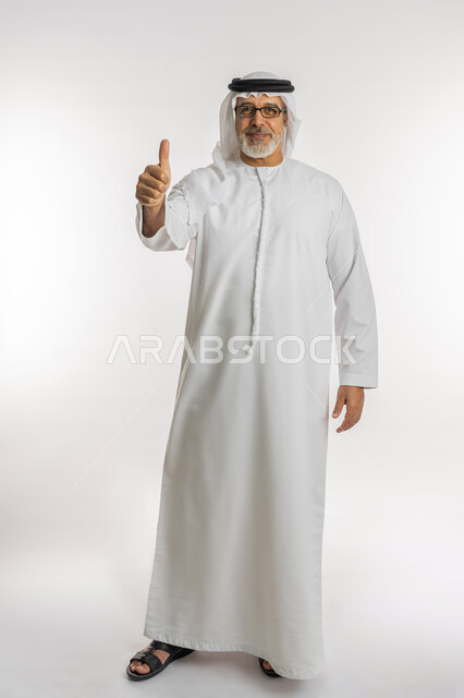 الشعور بالرضا و القبول, علامة الجودة والامتياز, بورتريه لرجل كبير السن عربي خليجي اماراتي يرتدي الكندورة و الغترة والنظارات الطبية يرفع اصبع الابهام للاعلى بإيماءات الموافقة, الاعجاب لتحقيق أمر معين, الجسم بالطول الكامل, خلفية بيضاء