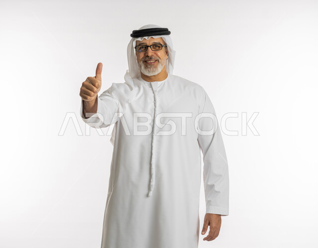 علامة الجودة والامتياز, بورتريه لرجل كبير السن عربي خليجي اماراتي يرتدي الكندورة و الغترة والنظارات الطبية يرفع اصبع الابهام للاعلى بإيماءات الموافقة, الاعجاب لتحقيق أمر معين, الشعور بالرضا و القبول, خلفية بيضاء