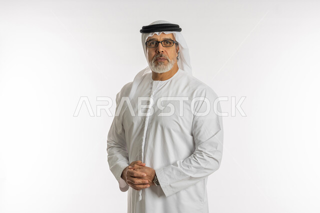 مفهوم الاهتمام بالمظهر الخارجي و الرجولة والأناقه، بورتريه مقرب لرجل مسن عربي خليجي اماراتي يرتدي الكندورة و الغترة يقف باستقامة بايماءات ايدي متماسكة ,النظر إلى الكاميرا يإيماءات العبوس، تعابير الجدية والثقة بالنفس، صورة الجسم بالطول الكامل، خلفية بيضاء