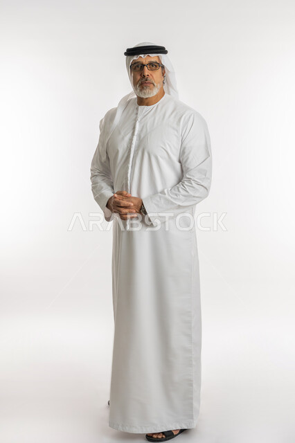 تعابير الجدية والثقة بالنفس، مفهوم الاهتمام بالمظهر الخارجي و الرجولة والأناقه، بورتريه لرجل مسن عربي خليجي اماراتي يرتدي الكندورة و الغترة يقف باستقامة بايماءات ايدي متماسكة ,النظر إلى الكاميرا يإيماءات العبوس، صورة الجسم بالطول الكامل، خلفية بيضاء