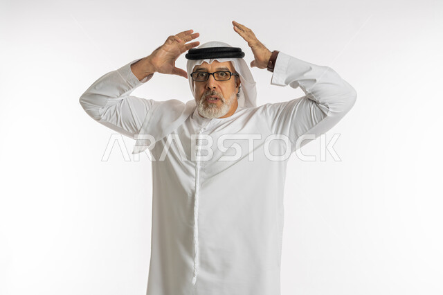 الصدمة و المفاجأة برؤية شيء ما، الانزعاج عند سماع خبر سيء ,الوقوع في مصيبة, النظر الى الكاميرا بتعابير التعجب, بورتريه مقرب لرجل عربي اماراتي خليجي يرتدي الكندورة و الغترة يضع يديه على رأس بتعابير الدهشة و الذهول، خلفية بيضاء