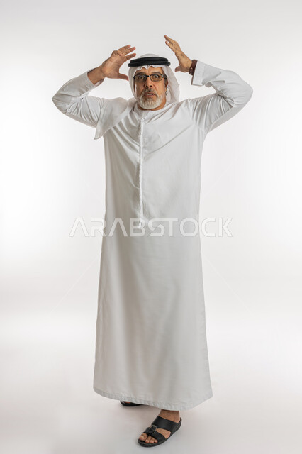 النظر الى الكاميرا بتعابير التعجب, الانزعاج عند سماع خبر سيء ,الوقوع في مصيبة, الصدمة و المفاجأة برؤية شيء ما، بورتريه لرجل عربي اماراتي خليجي يرتدي الكندورة و الغترة يضع يديه على رأس بتعابير الدهشة و الذهول، صورة الجسم بالطول الكامل، خلفية بيضاء