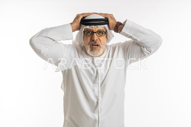 المفاجأة و الصدمة عند رؤية شيء ما، الانزعاج عند سماع خبر سيء ,الوقوع في مصيبة, النظر الى الكاميرا بتعابير التعجب, بورتريه مقرب لرجل عربي اماراتي خليجي يرتدي الكندورة و الغترة يضع يديه على رأس بتعابير الدهشة و الذهول، خلفية بيضاء