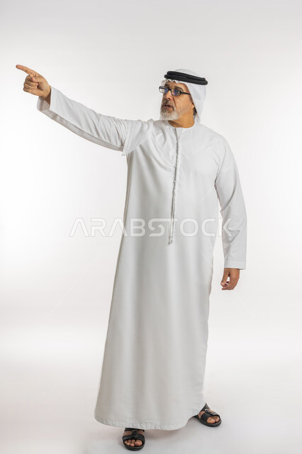 التحديد نحو هدف معين, بورتريه لرجل مسن عربي اماراتي خليجي يرتدي الكندورة و الغترة و النظارات الطبية يرفع اصبع السبابة للاعلى بايماءات التأكد من شيء ما, الإشارة الى شخص ما, صورة الجسم بالطول الكامل, خلفية بيضاء
