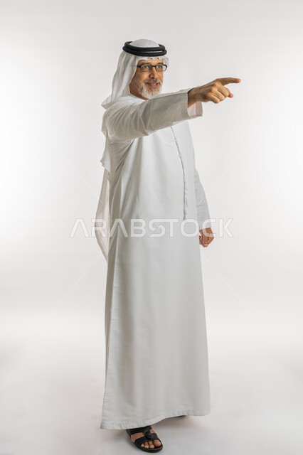 ايماءات التأكد من شيء ما ,التحديد نحو هدف معين, بورتريه لرجل مسن عربي اماراتي خليجي يرتدي الكندورة و الغترة و النظارات الطبية يرفع اصبع السبابة للاعلى بتعابير السرور, الإشارة الى شخص ما, صورة الجسم بالطول الكامل, خلفية بيضاء