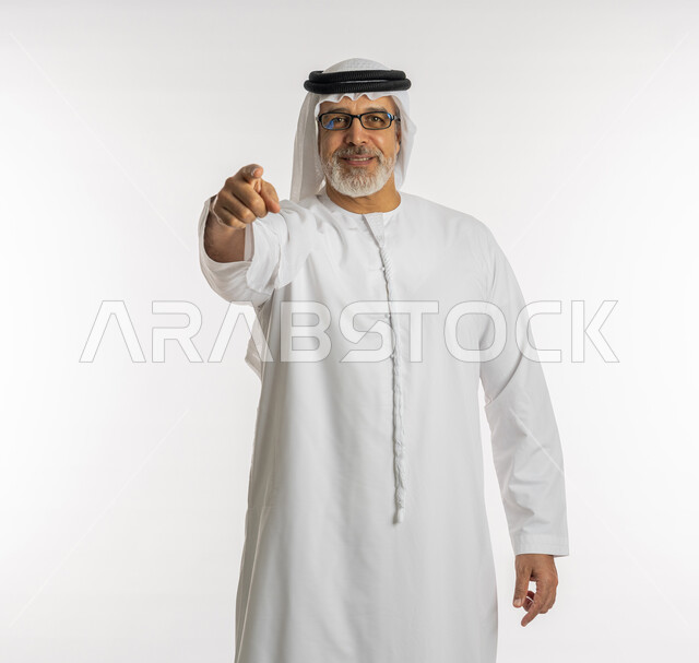 الإشارة الى شخص ما, النظر الى الكاميرا بايماءات التأكد ,التحديد نحو هدف معين, بورتريه لرجل مسن عربي اماراتي خليجي يرتدي الكندورة و الغترة و النظارات الطبية يرفع اصبع السبابة للاعلى بتعابير السرور, خلفية بيضاء