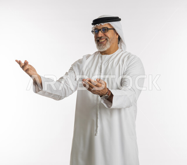 حركات تدل على الاستقبال و الترحيب, الاشارة الى شيء ما بايماءات السعادة والفرح, بورتريه مقرب لرجل مسن عربي اماراتي خليجي يرتدي الكندورة و الغترة يرفع ذراعيه للاعلى بتعابير الضحك ,خلفية بيضاء