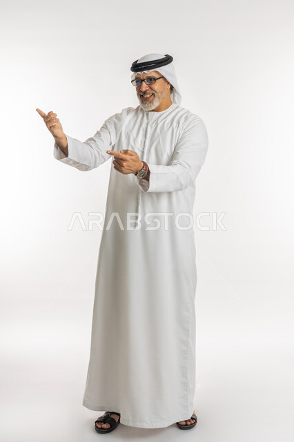 الاشارة الى شيء ما بايماءات السعادة والفرح, بورتريه لرجل مسن عربي اماراتي خليجي يرتدي الكندورة و الغترة يرفع ذراعيه للاعلى بتعابير الضحك ,قضاء وقت ممتع في العطلة, صورة الجسم بالطول الكامل, خلفية بيضاء
