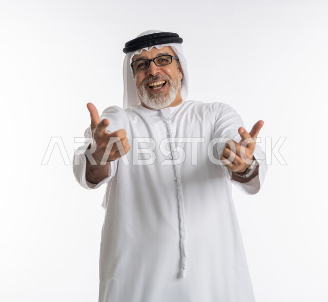 ايماءات السعادة والفرح, بورتريه مقرب لرجل مسن عربي اماراتي خليجي يرتدي الكندورة و الغترة يرفع ذراعيه للاعلى بتعابير الضحك الهستيري, النظر الى الكاميرا بحركات السرور ,قضاء وقت ممتع في العطلة, خلفية بيضاء