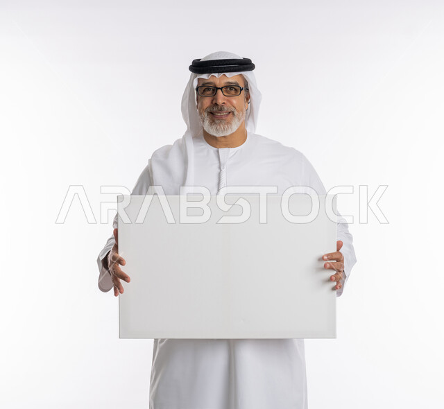 الوقوف باستقامه وثقه، لوح إعلاني فارغ، إيماءات الإعجاب والسرور والنظر إلى الكاميرا ،موك اب مستطيل أبيض ، بورتريه لرجل مسن عربي اماراتي خليجي يرتدي الكندورة و الغترة يحمل بيديه لوحه بيضاء مستطيلة، خلفية بيضاء